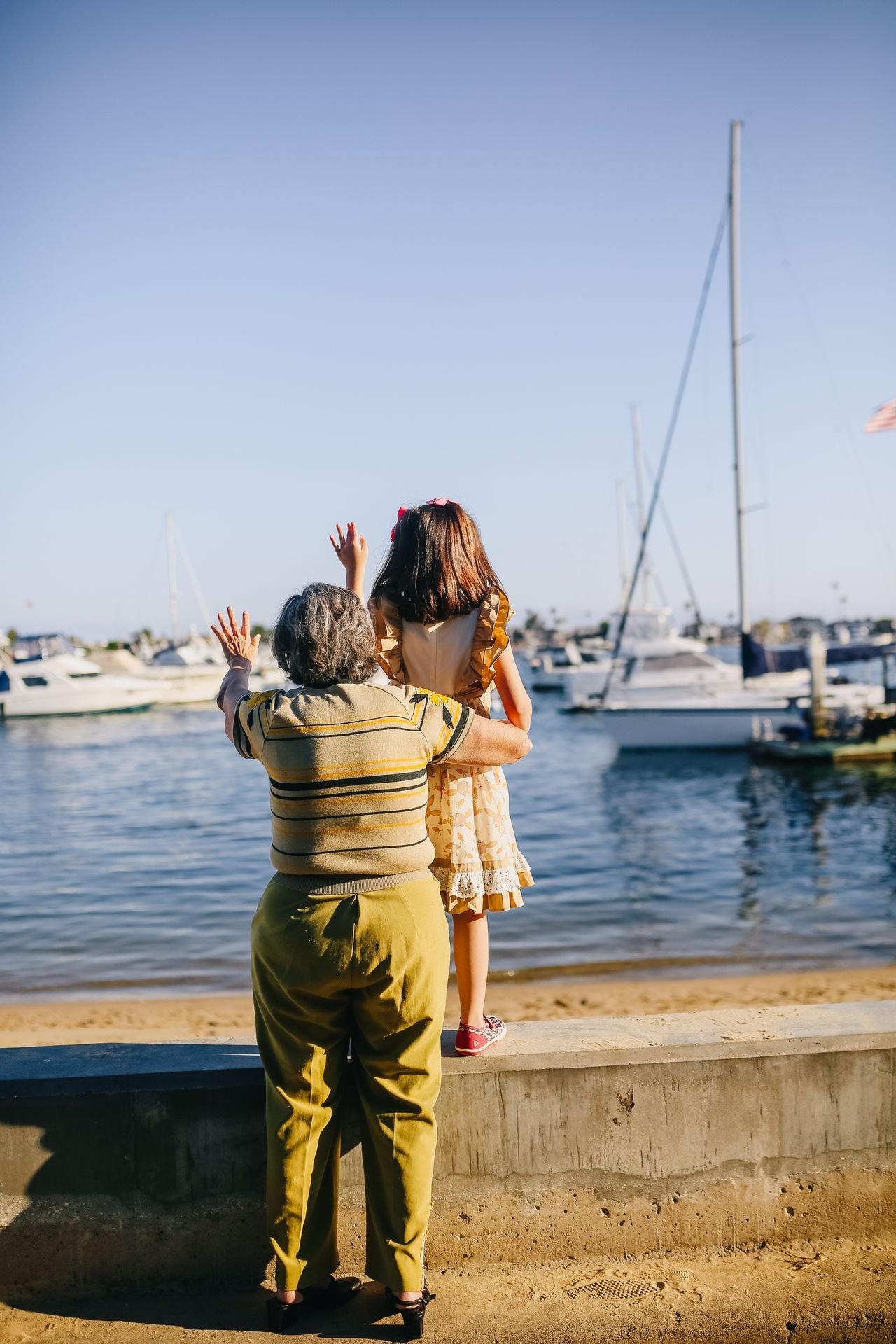 A grandmother and her granddaughter looking out at the boats in a harbor, seen from behind