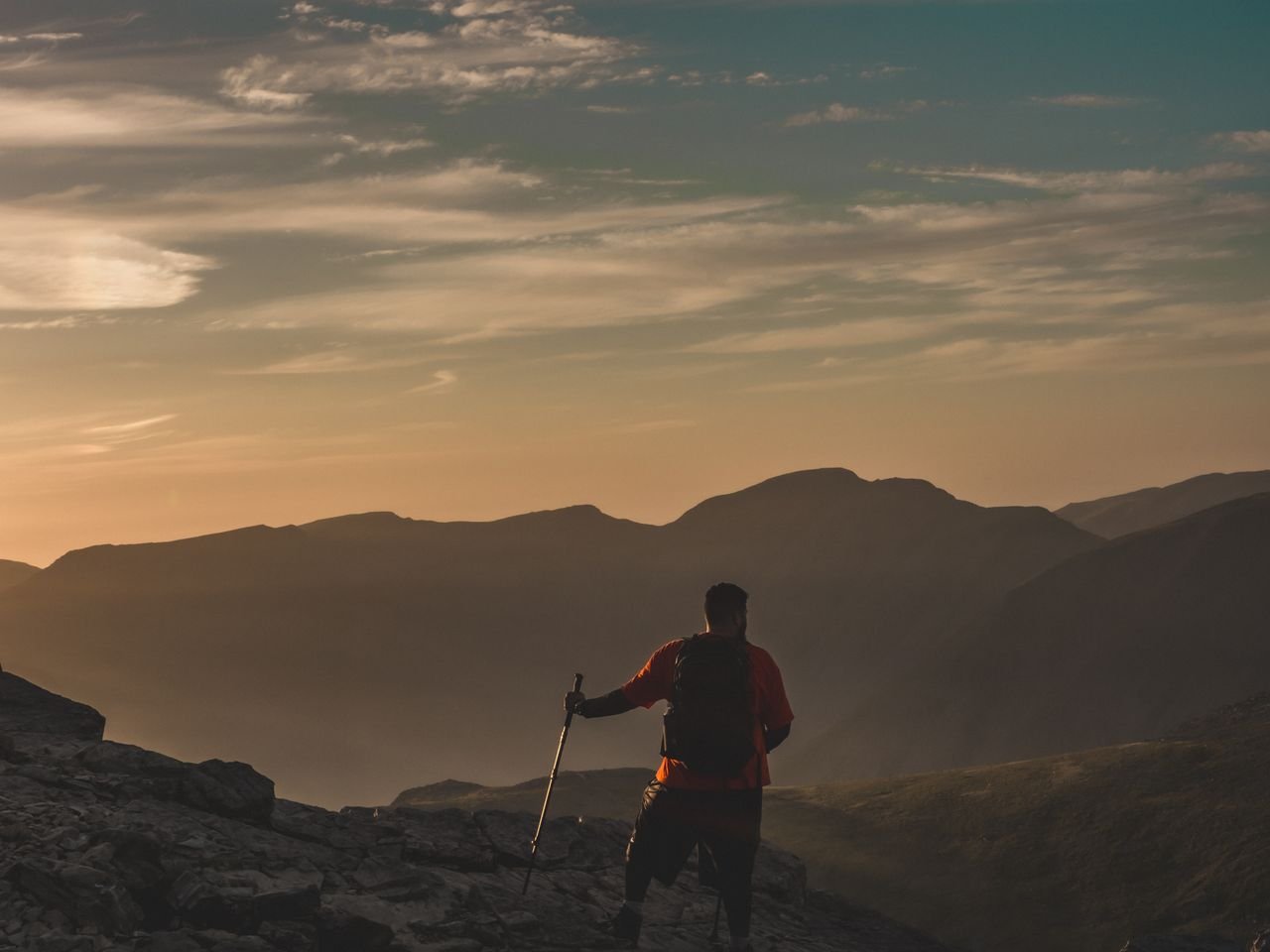A hiker with a walking pole looking out over the mountains at sunset