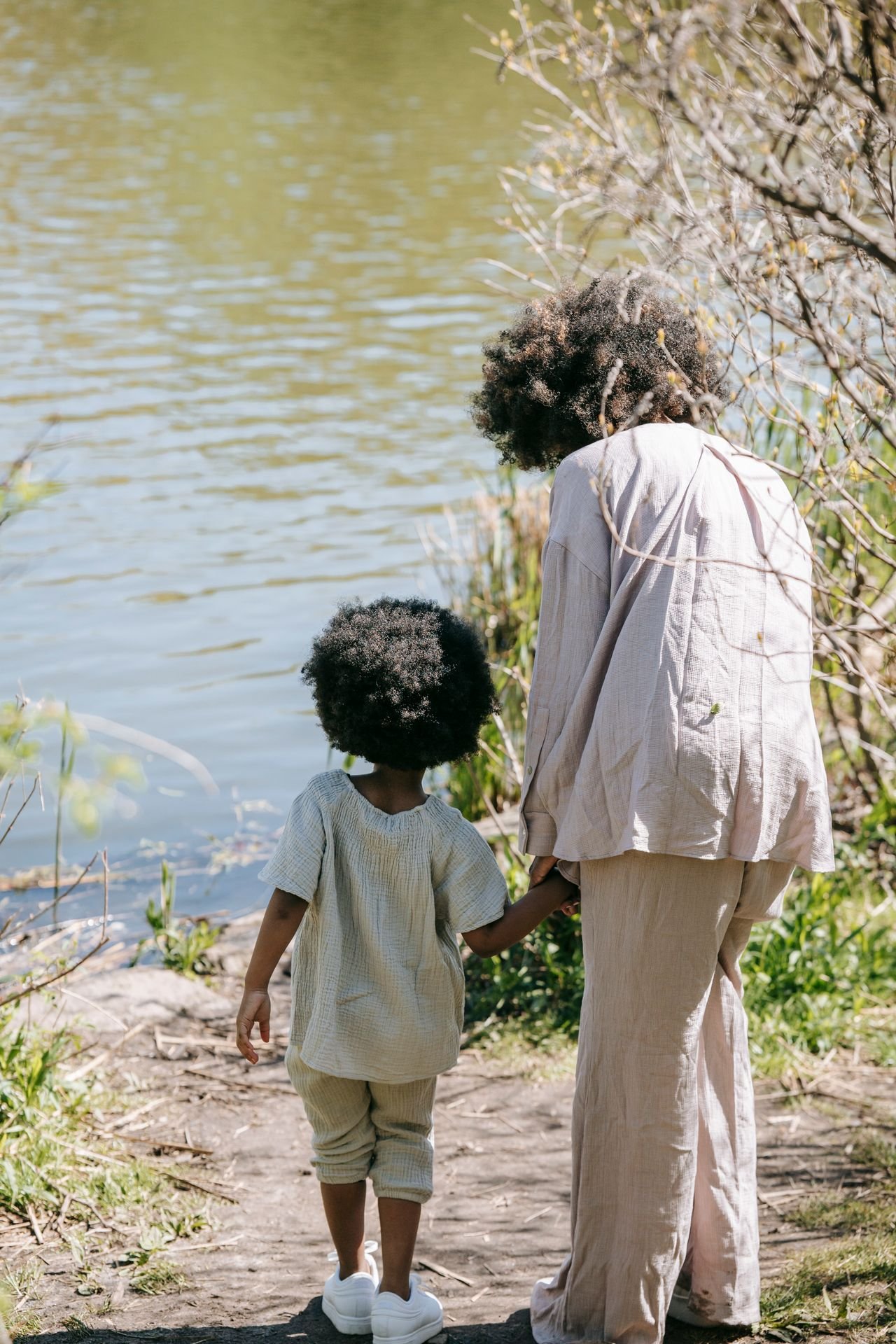 A mother and her young child standing hand in hand by a lake, seen from behind
