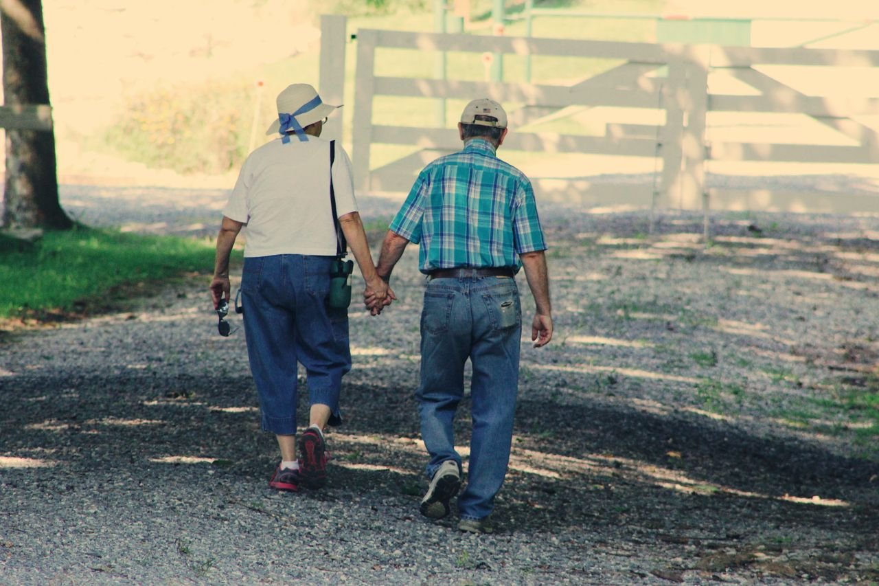 An older couple walking hand in hand down a sunny country path, seen from behind