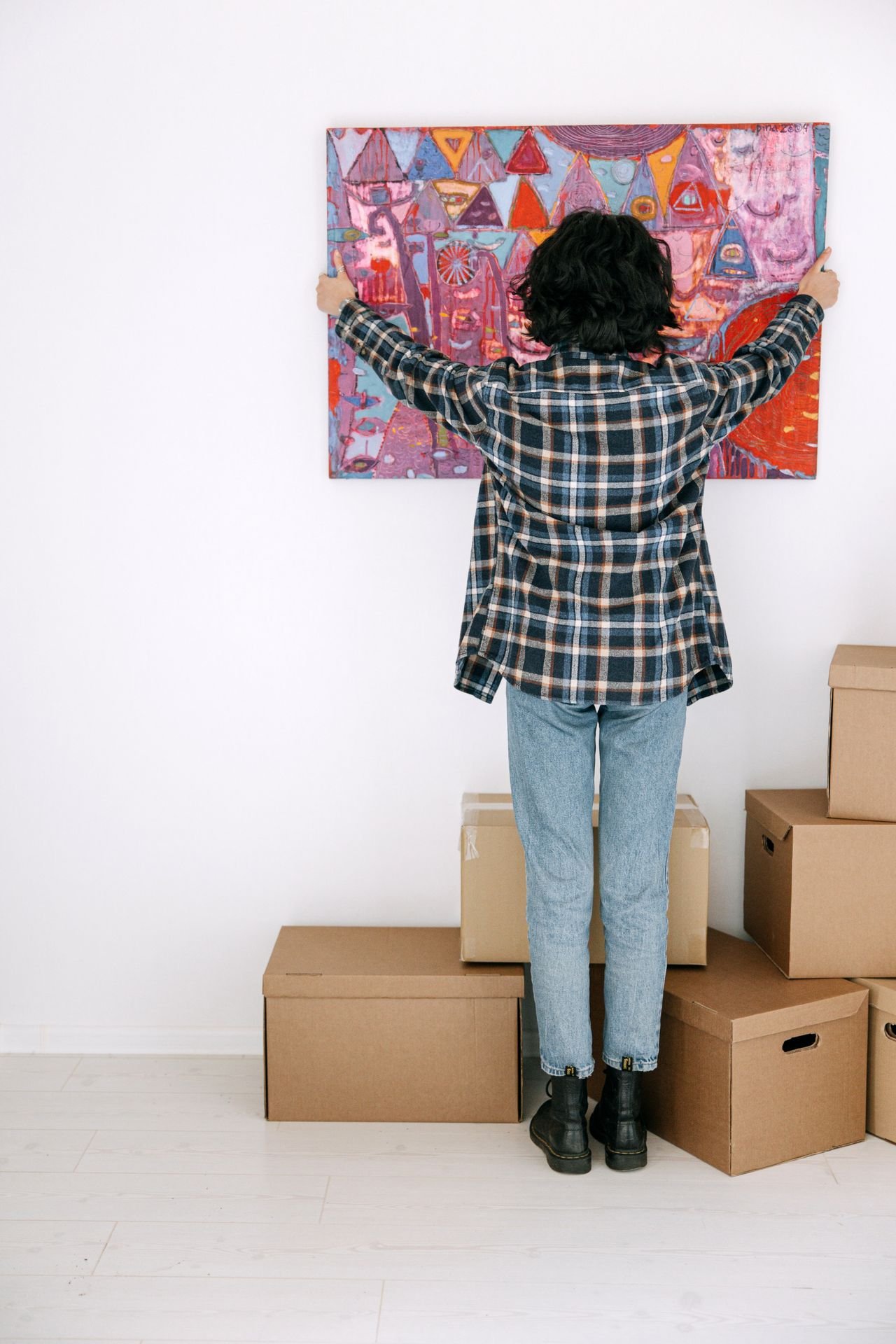 A woman hanging a colorful painting in her new apartment among moving boxes, seen from behind